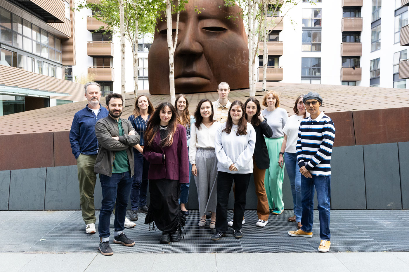 A group of 12 people stand together outside in front of a statue on a clear and sunny day