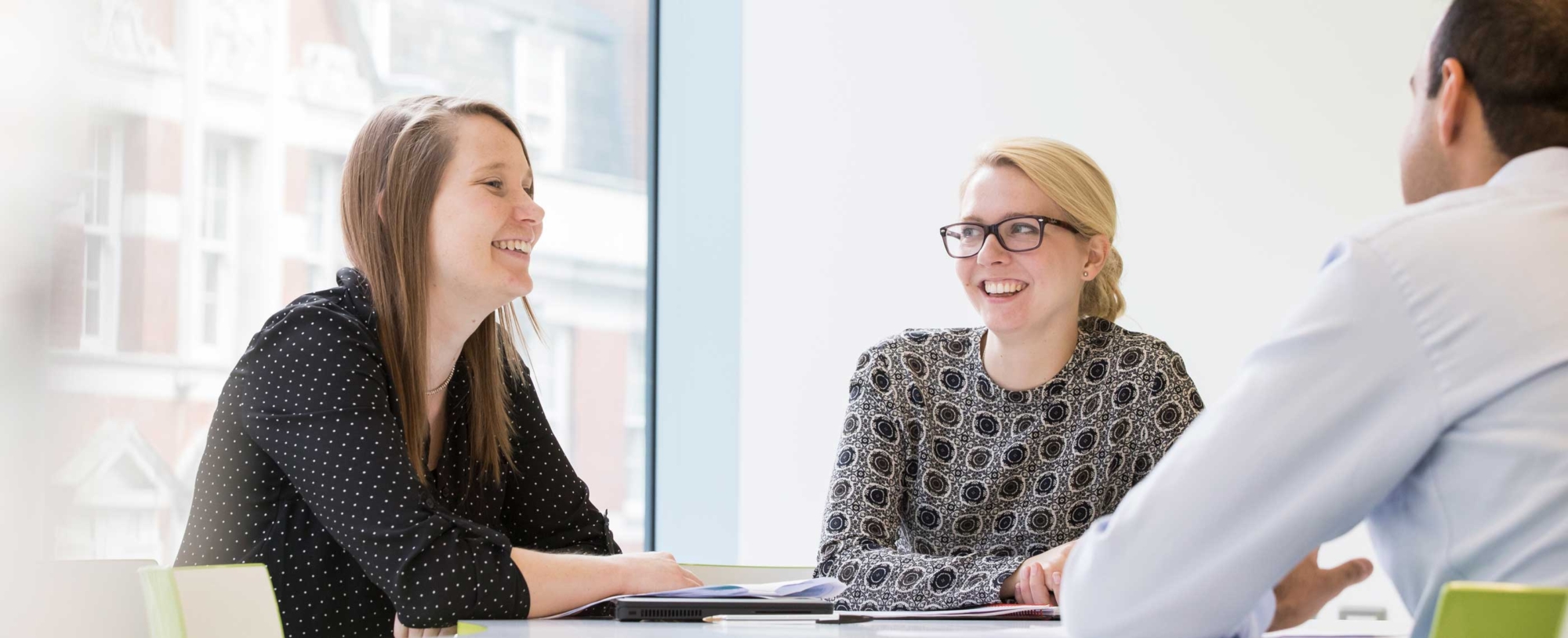 A group of office workers sit at a table in front of a large window talking to each other and smiling