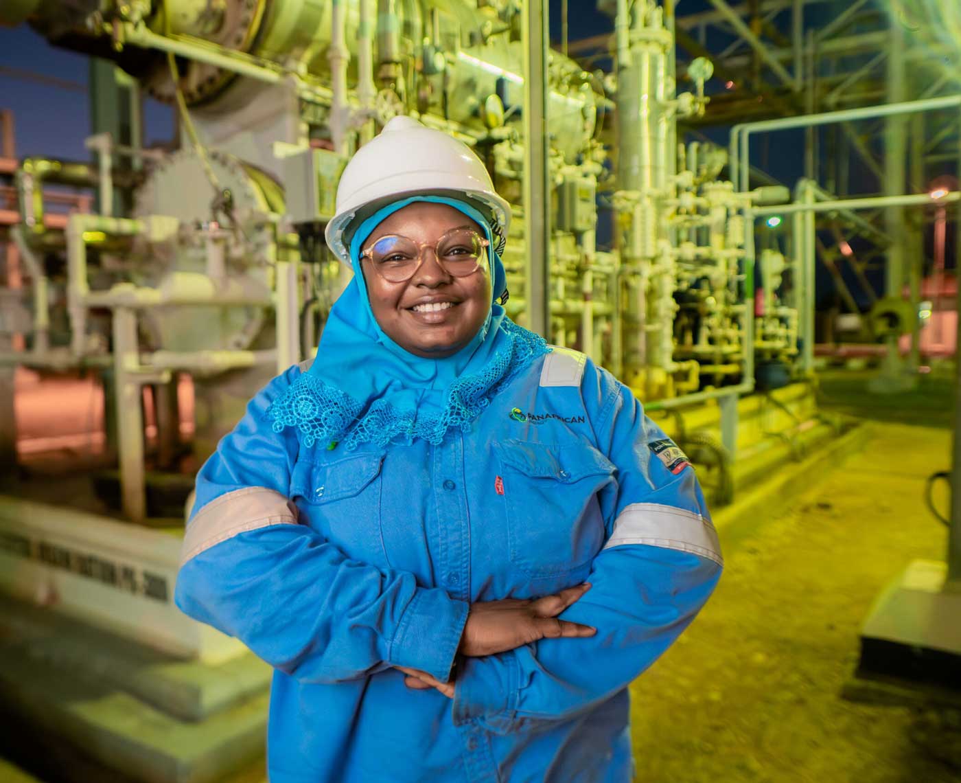 A smiling woman wearing blue coveralls, a hard hat and safety googles stands in a gas rig at night