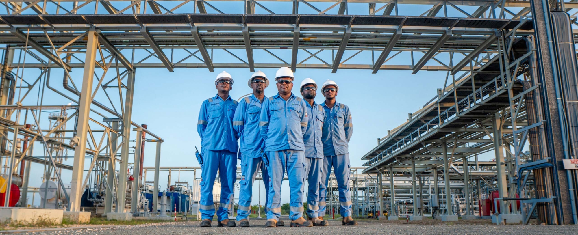 5 men stand as a group in front of a gas rig, they are wearing blue coveralls and safety equipment with the PAET brand on
