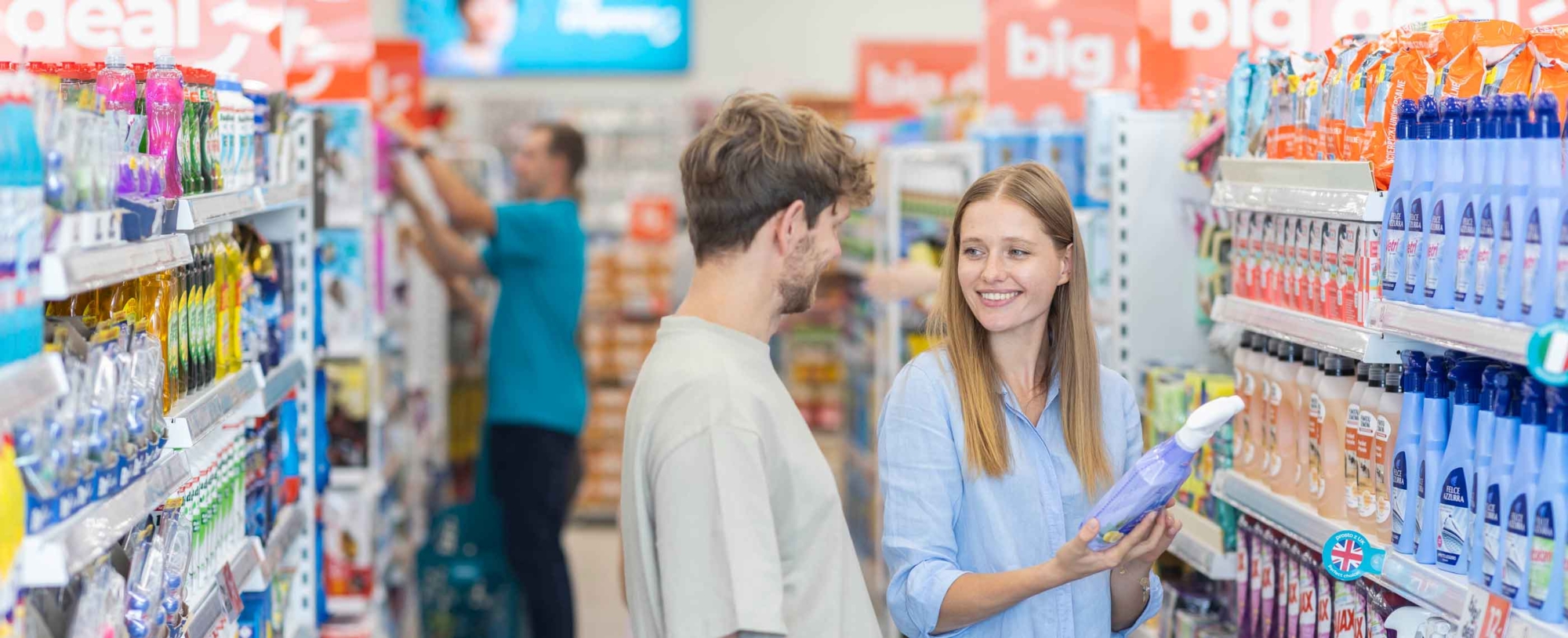 A man and a smiling woman stand in the isle of a supermarket, with an employee stacking shelves in the background