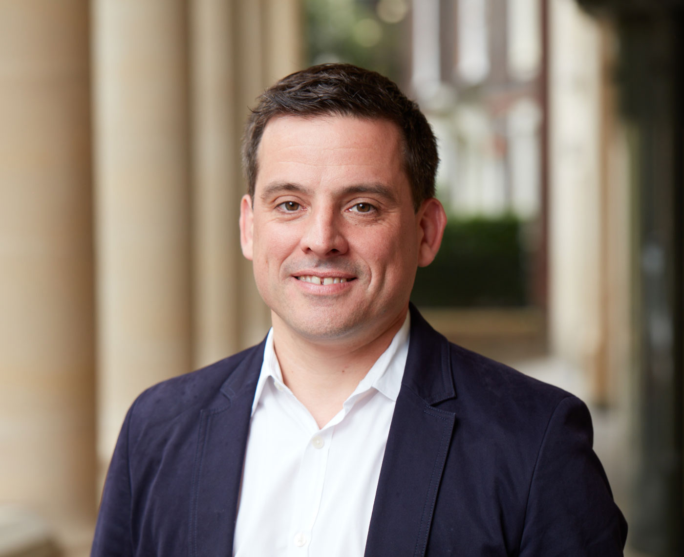 A headshot of a man standing outside and looking at camera wearing a suit and open collar